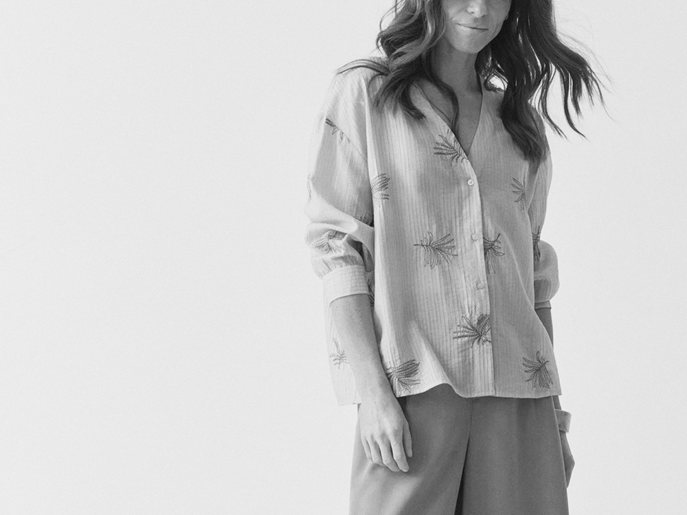 O femeie îmbrăcată într-o cămașă bej cu imprimeu floral în nuanțe de albastru și verde privește în jos cu un zâmbet blând.