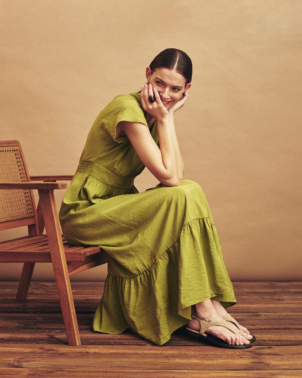 Žena v dlouhých zelených šatech, sedící na dřevěné židli, usmívá se a opírá si tvář o ruce.
